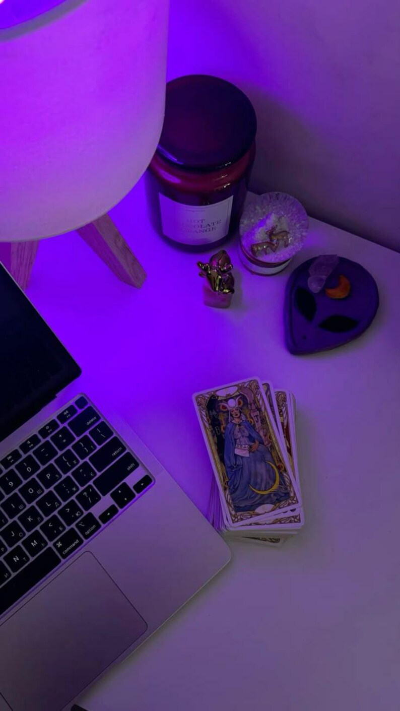 May include: A laptop, tarot cards, and decorative items are arranged on a white surface, illuminated by a purple light. The tarot cards feature illustrations of figures in ornate clothing. Other items include a candle, a small gold statue, and a purple alien-shaped dish.
