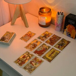 May include: A white table displays a tarot card reading setup. Tarot cards with gold and yellow illustrations are spread out. A lit candle, a lamp, and a Zodiac tarot card box are also on the table.
