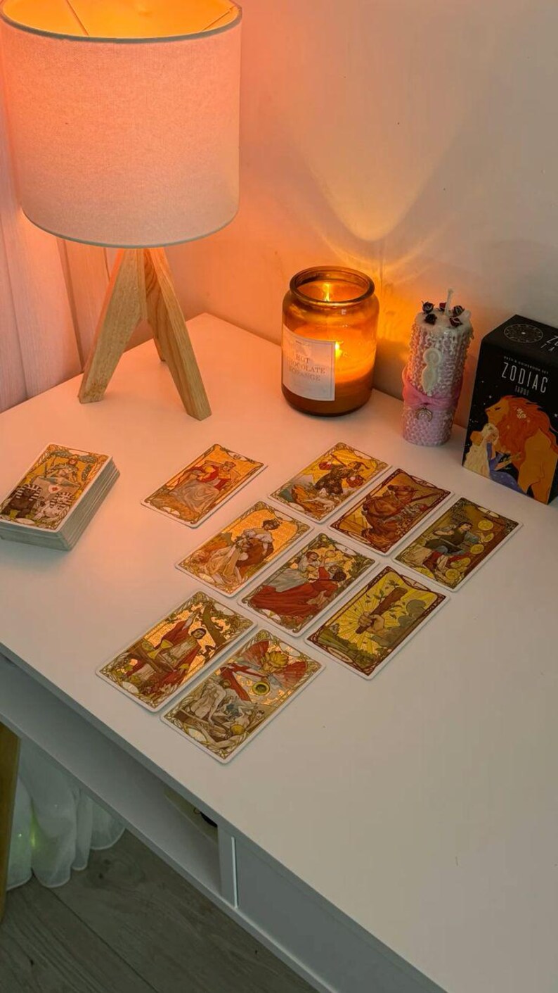 May include: A white table displays a tarot card reading setup. Tarot cards with gold and yellow illustrations are spread out. A lit candle, a lamp, and a Zodiac tarot card box are also on the table.