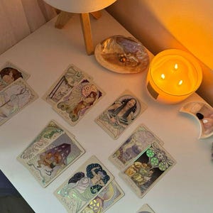 May include: A collection of tarot cards with Art Nouveau-style illustrations, featuring figures and symbols. The cards are arranged on a white surface, alongside a lit candle, a decorative shell, and a small lamp.