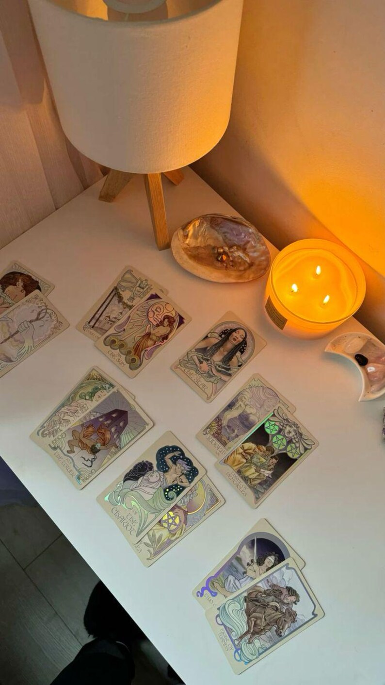 May include: A collection of tarot cards with Art Nouveau-style illustrations, featuring figures and symbols. The cards are arranged on a white surface, alongside a lit candle, a decorative shell, and a small lamp.