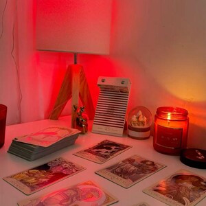 May include: A well-lit scene featuring tarot cards, a lit candle, and a lamp. The tarot cards are spread out, with a stack of cards to the left. A small snow globe and a calendar are also present. The scene is illuminated by a warm, red light.