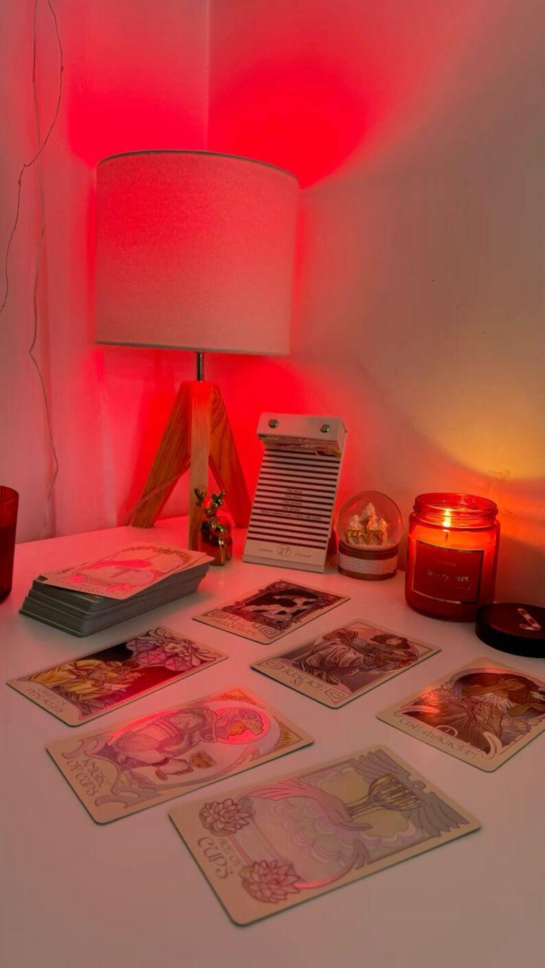 May include: A well-lit scene featuring tarot cards, a lit candle, and a lamp. The tarot cards are spread out, with a stack of cards to the left. A small snow globe and a calendar are also present. The scene is illuminated by a warm, red light.
