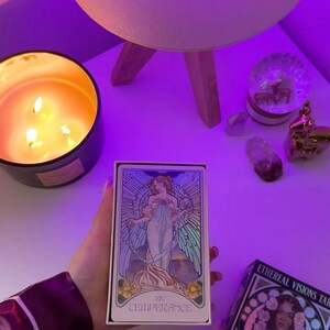 May include: A tarot card box and deck featuring the Temperance card, held near a lit candle and a lamp. The card art depicts a woman in flowing robes. The box reads "XIV TEMPERANCE."