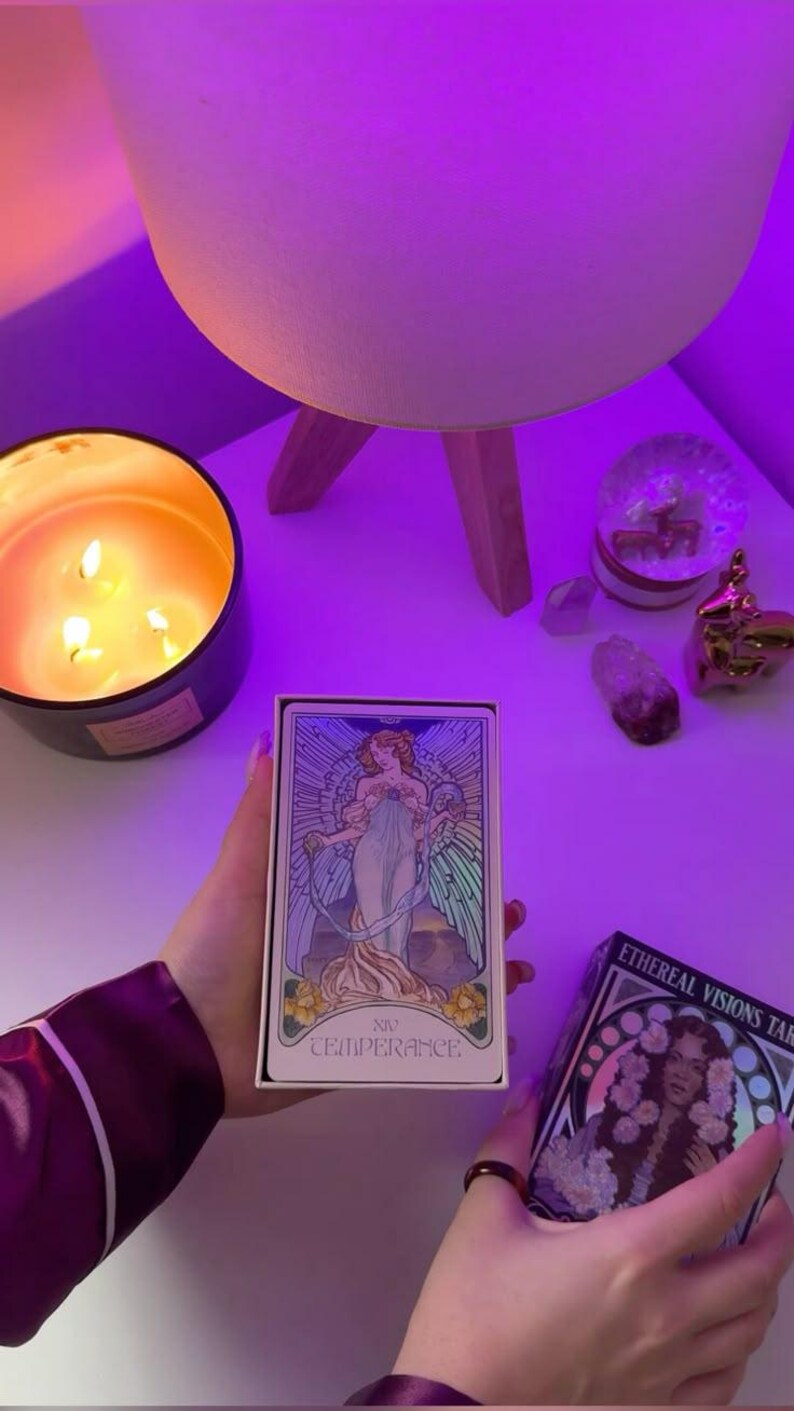 May include: A tarot card box and deck featuring the Temperance card, held near a lit candle and a lamp. The card art depicts a woman in flowing robes. The box reads "XIV TEMPERANCE."