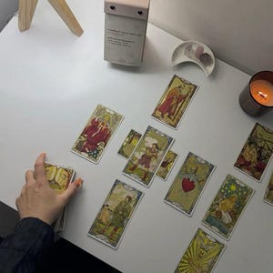 May include: A tarot card reading setup on a white surface. Several tarot cards are spread out, featuring colorful illustrations. A lit candle, a small decorative dish, and a small calendar are also present.
