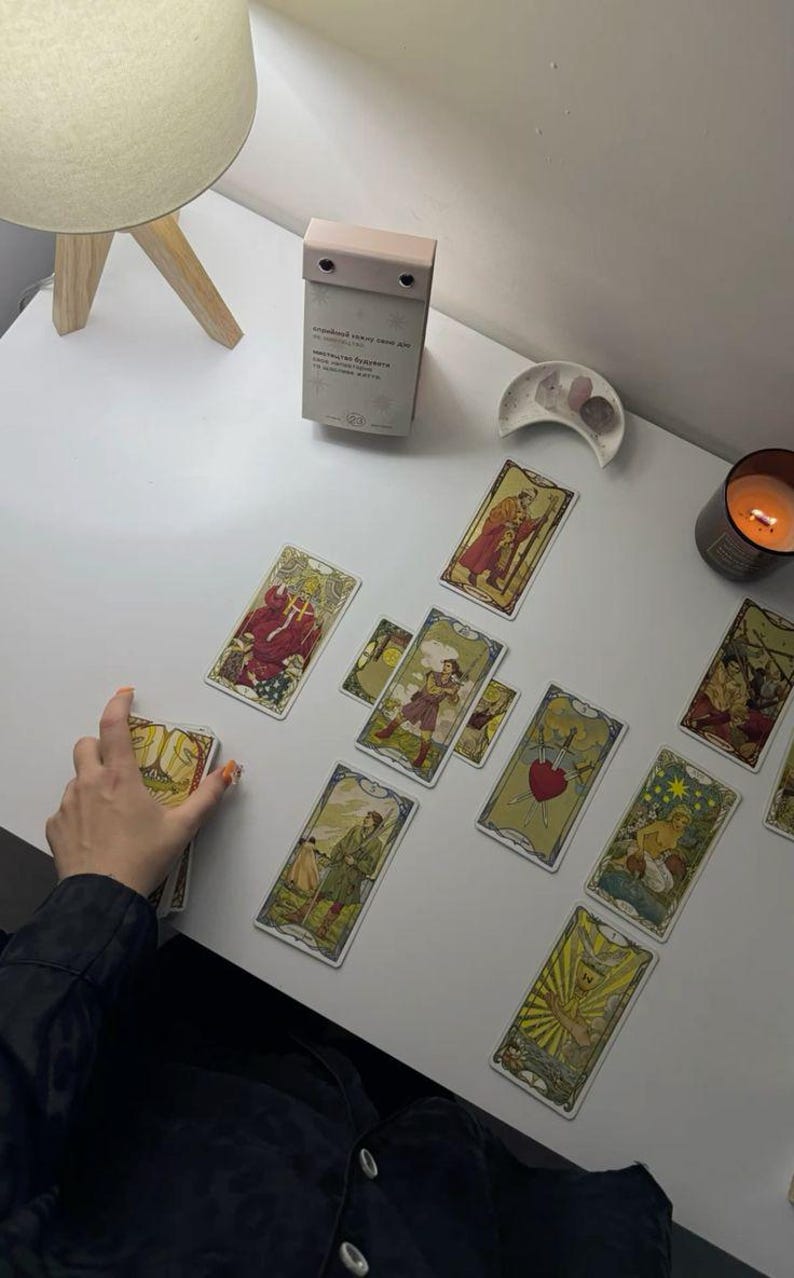 May include: A tarot card reading setup on a white surface. Several tarot cards are spread out, featuring colorful illustrations. A lit candle, a small decorative dish, and a small calendar are also present.