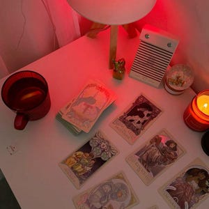 May include: A collection of tarot cards with illustrations, arranged on a white surface. A red mug, a candle, and a small snow globe are also present. The scene is illuminated by a red-toned lamp.