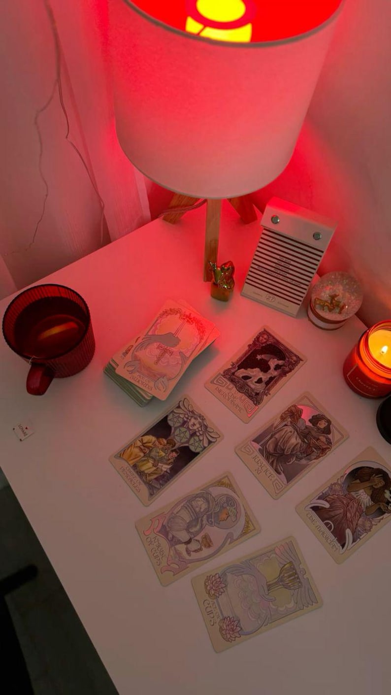 May include: A collection of tarot cards with illustrations, arranged on a white surface. A red mug, a candle, and a small snow globe are also present. The scene is illuminated by a red-toned lamp.
