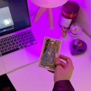 May include: A tarot card featuring a woman in a blue dress and a crescent moon, held in a hand with an orange ring. The card is on a white surface, with a laptop, lamp, and other items in the background. The scene is illuminated with a purple light.