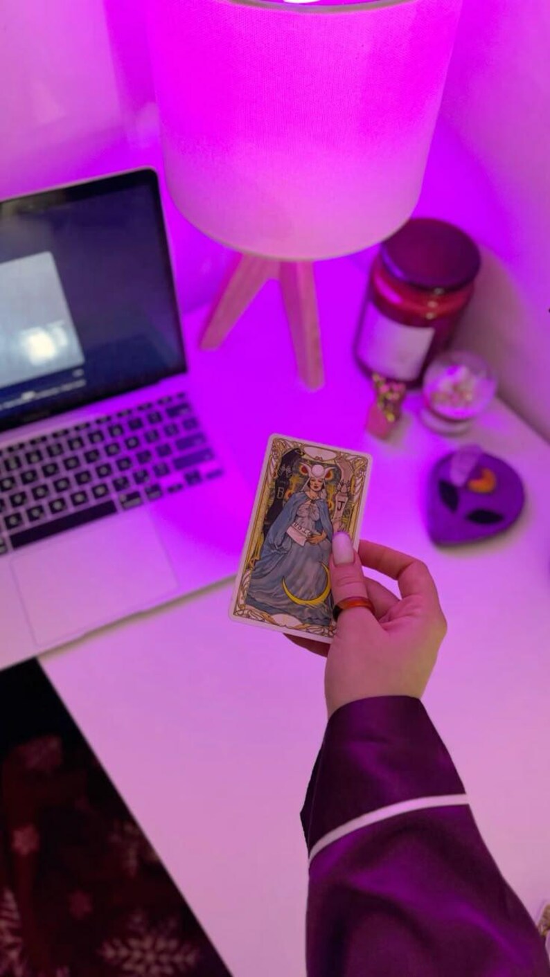 May include: A tarot card featuring a woman in a blue dress and a crescent moon, held in a hand with an orange ring. The card is on a white surface, with a laptop, lamp, and other items in the background. The scene is illuminated with a purple light.