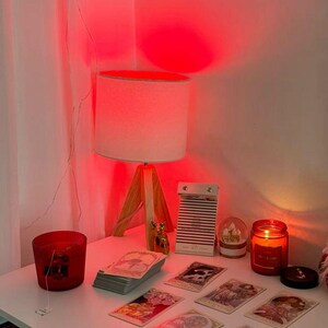 May include: A white table with a wooden-legged lamp, a red candle, and a stack of tarot cards. Other items include a small snow globe, a decorative deer figurine, and a small notebook. The scene is illuminated by a red light.