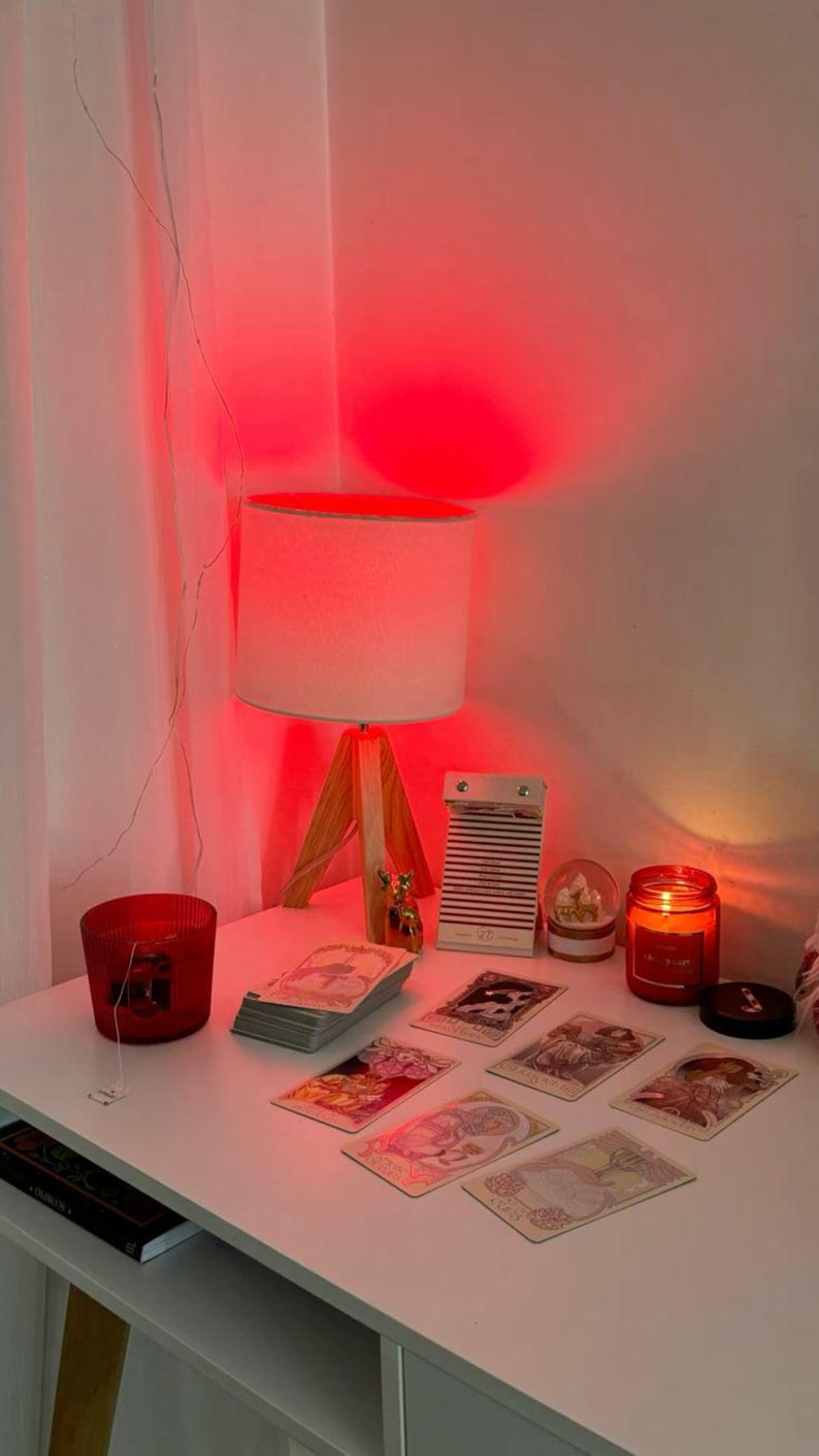 May include: A white table with a wooden-legged lamp, a red candle, and a stack of tarot cards. Other items include a small snow globe, a decorative deer figurine, and a small notebook. The scene is illuminated by a red light.