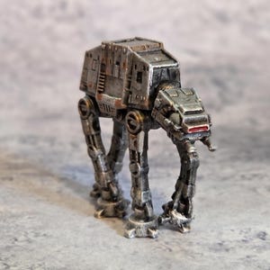 Puede incluir: Modelo detallado y desgastado de un caminante AT-AT de Star Wars. El vehículo gris y plateado tiene cuatro patas, un cuerpo en forma de caja y una cabeza con un acento rojo. El modelo está sobre una superficie gris.