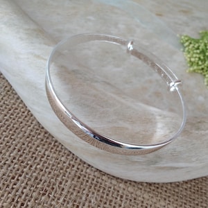 May include: A polished silver-tone bangle bracelet featuring a smooth, curved surface and an adjustable sliding mechanism with a small metal bead, designed to expand or contract for a comfortable fit on the wrist.