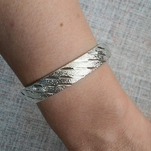 May include: A silver-toned metal bangle bracelet featuring a textured surface with diagonal, raised ridges. The bracelet has a polished finish that reflects light, creating a repeating pattern of metallic lines across the entire outer band.