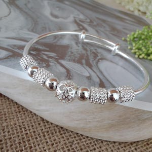May include: A silver-toned expandable bangle bracelet featuring a series of alternating beads. The design includes smooth round beads, textured cylindrical beads with a sparkling finish, and a central spherical bead with a cut-out heart pattern.