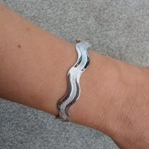 May include: A silver-toned metal bangle bracelet featuring a distinct wavy, serpentine silhouette. The surface of the bracelet displays a contrasting design with smooth, polished edges and a central band characterised by a fine, cross-hatched texture.