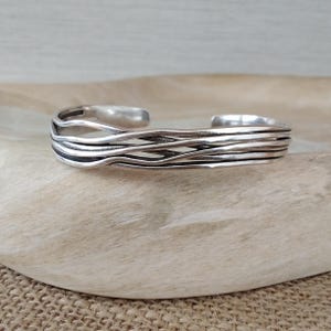 May include: A silver cuff bracelet featuring multiple thin, overlapping, and wavy metal strands that create a layered, open-work design. The polished metal surface reflects light, highlighting the fluid, horizontal lines of the band.
