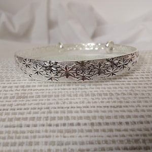 May include: A silver bangle bracelet featuring a repeating starburst design around the band. The bracelet is adjustable and rests on a white textured surface. The metal has a polished finish and the design is detailed.