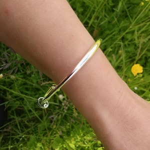 May include: A polished silver-tone bangle bracelet featuring a smooth, rounded band. The bracelet has a circular clasp mechanism at one end, which is accented with a small, clear, round gemstone that catches the light, creating a subtle sparkle.