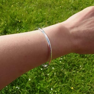 May include: A polished silver-tone bangle bracelet with a smooth, rounded surface. The bracelet features a simple, continuous circular design that rests comfortably on the wrist, showcasing a reflective metallic finish and a minimalist aesthetic.