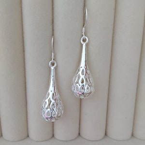 May include: A pair of silver-toned drop earrings featuring a teardrop-shaped pendant with an intricate, openwork lattice design. Each earring hangs from a simple silver hook, showcasing a polished metallic finish and a hollow, cage-like structure.