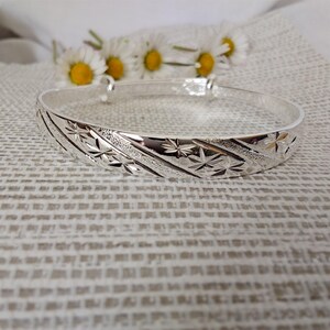 May include: A silver adjustable bangle bracelet with a starburst design. The bracelet has a polished finish and is set against a textured background with white flowers.