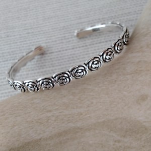 May include: A silver-toned cuff bracelet featuring a continuous row of embossed rose motifs. The metal has a polished finish with darkened recessed areas to highlight the floral details. The bracelet has an open design with rounded ends.