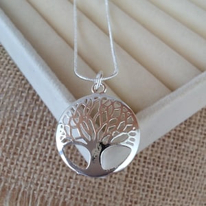 May include: A silver-coloured necklace featuring a circular pendant with a cut-out tree of life design. The pendant is suspended from a thin, flexible silver-coloured snake chain. The metal has a polished, reflective finish that catches the light.