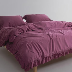 May include: A bed with a plum-colored duvet cover and matching pillowcases. The duvet cover and pillowcases have a ruffled edge. The bed frame is made of light-colored wood.