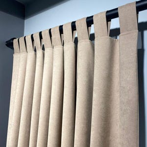 May include: Beige fabric curtains hanging from a black rod. The curtains have a textured appearance and are suspended by fabric loops. The background is a light blue wall.
