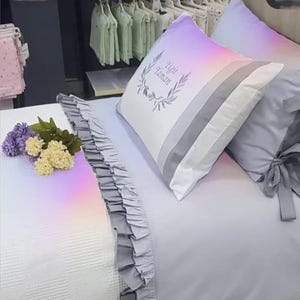 May include: A bed set featuring a white comforter with a gray ruffled edge and a gray pillowcase with a bow. A pillow with a gradient of colors and the text "Yigit Yaman" is also visible. The set includes a bouquet of purple and yellow flowers.