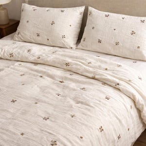 May include: Off-white bedding set with embroidered floral accents. The set includes a duvet cover and two pillowcases. The fabric has a textured appearance, and the floral embroidery is in a muted brown color. The bedding is arranged on a bed with a neutral-toned headboard.