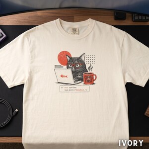 Op de afbeelding: Ivoor kleurig t-shirt met een afbeelding van een zwarte kat met een bril, werkend op een laptop met een visgraatmotief. Een rode mok met "COFFEE FIRST" staat naast de kat. De tekst "If not coffee: sys.exit("Goodbye.")" is inbegrepen.