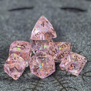 Puede incluir: Un juego de siete dados poliédricos hechos de resina rosa translúcida con escamas de papel iridiscente incrustadas que captan la luz. Cada dado presenta bordes afilados y números grabados en color oscuro para facilitar su lectura.