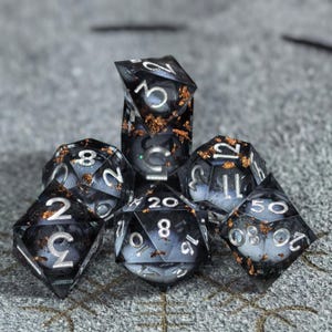 May include: A set of seven polyhedral dice made of translucent dark gray resin with embedded copper-colored metallic flakes. Each die features sharp edges and clear, silver-colored numbers engraved on the faces for high visibility and contrast.