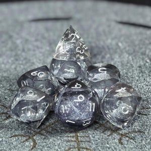 Könnte beinhalten: Ein Set aus sieben polyedrischen Würfeln aus klarem Harz mit silbernem Glitzer im Inneren. Jeder Würfel hat scharfe Kanten und weiß gravierte Zahlen. Das Set enthält einen W4, W6, W8, W10, W12, W20 und einen Prozentwürfel, die zusammen angeordnet sind.