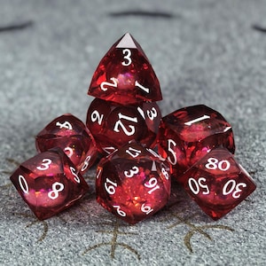 Puede incluir: Un juego de siete dados poliédricos de color rojo oscuro translúcido con números blancos. Los dados contienen partículas de purpurina iridiscente incrustadas que captan la luz. El set incluye un d4, d6, d8, d10, d12, d20 y un dado porcentual.