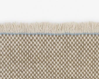 May include: A woven textile featuring a tight, repeating checkerboard pattern in alternating shades of tan and off-white. The top edge is finished with a thin, horizontal light blue thread line and a row of short, cream-colored fringe.