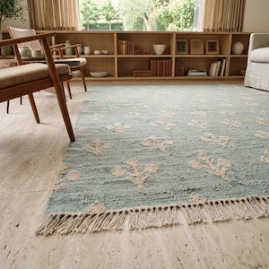 May include: A rectangular area rug featuring a light blue base with a repeating cream-colored floral pattern and a fringed edge. The rug has a textured pile surface and is positioned on a light-colored stone floor in a room with wooden furniture.