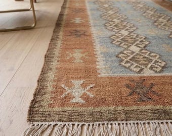 Puede incluir: Una alfombra rectangular que presenta un panel central azul claro con un patrón de diamantes marrones repetitivo, rodeado por un borde ancho de color terracota con motivos geométricos blancos y marrones oscuros, acabado con flecos blanquecinos.