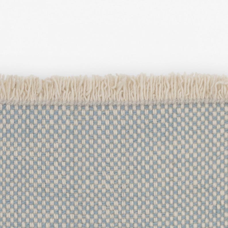 Pode incluir: Um tecido com um padr&atilde;o de grelha repetitivo de fios azul-claros e branco-sujo. A extremidade do tecido &eacute; rematada com uma fila de franjas curtas e densas de cor branco-sujo que se estendem horizontalmente pelo topo do material.
