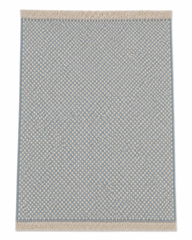Pode incluir: Um tapete retangular com um padr&atilde;o tecido de fios azul-claros e branco-sujo. O tapete apresenta um design de grelha denso e texturizado, e &eacute; rematado com franjas curtas e direitas de cor branco-sujo nas extremidades superior e inferior.