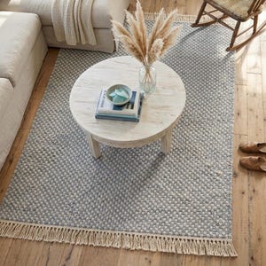 Pode incluir: Um tapete retangular com um padr&atilde;o xadrez azul e creme, uma trama texturizada e franjas longas nas bordas. O tapete est&aacute; colocado sobre um piso de madeira e serve de base para uma mesa de centro redonda de madeira branca com um vaso.