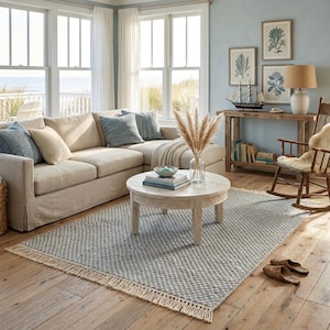 Pode incluir: Um tapete retangular com um padr&atilde;o texturizado tecido em azul e branco, com franjas atadas nas extremidades curtas. O tapete est&aacute; posicionado sobre um piso de madeira clara, parcialmente coberto por uma mesa de centro redonda e um sof&aacute;.