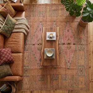 May include: A large rectangular area rug featuring a faded terracotta and pink palette with geometric patterns in muted green and yellow. The rug is positioned under a brown leather sofa with patterned throw pillows and a live-edge wooden coffee table.