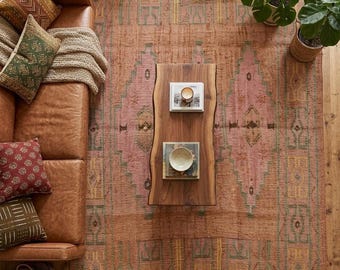 May include: A large rectangular area rug featuring a faded terracotta and pink palette with geometric patterns in muted green and yellow. The rug is positioned under a brown leather sofa with patterned throw pillows and a live-edge wooden coffee table.