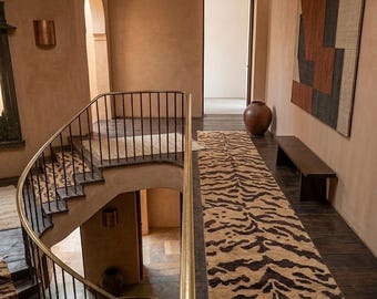 May include: A curved staircase with a brass handrail and black metal balusters features a runner with a brown and black tiger stripe pattern. A long matching runner lies on the floor next to a dark wooden bench and a large brown ceramic vase.