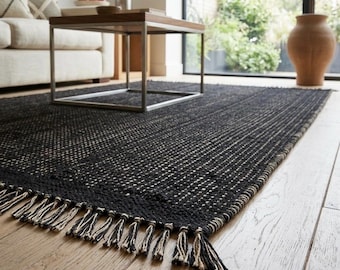 May include: A rectangular area rug featuring a dark black and off-white woven texture with a grid-like pattern. The rug is finished with short, knotted fringe tassels along the edge, creating a tactile surface that covers the wooden floor.